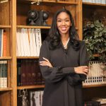Kyra Harris Bolden stands in front of a bookcase wearing black with arms folded